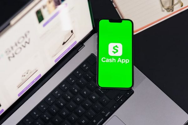 Smartphone showing Cash App screen on laptop keyboard, next to glasses and notebook.