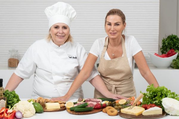 Two female chefs with variety of fresh ingredients in a kitchen setting, showcasing culinary preparation.